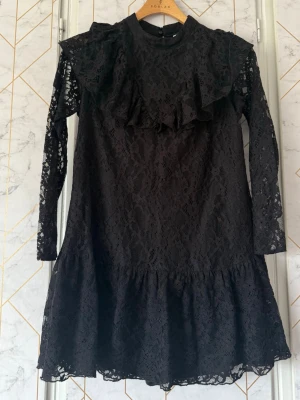 Svart spetsklänning med volang - Svart kort klänning från Zara i spets med långärmad design och volangdetaljer både fram och bak. Hög hals och lätt utställd kjol ger en trendig siluett. Med inbyggda short