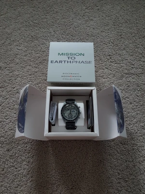 Omega x Swatch Mission to Earthphase - Snygg Omega x Swatch Mission to Earthphase från Bioceramic MoonSwatch Collection. Klockan har svart och silverfärgat boett, svart textilband med Omega-logga och tydlig tachymeter-skala på bezeln. Levereras i originalbox med manual och certifikat.