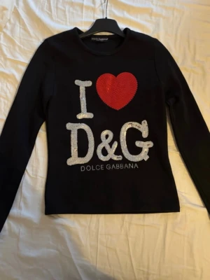 Svart paljettopp från Dolce & Gabbana - Svart långärmad topp från Dolce & Gabbana med stora paljetter framtill. Texten 'I ❤️ D&G' pryder bröstet med ett rött hjärta och silvriga bokstäver. Rund halsringning och figurnära passform. Perfekt statement-plagg för dig som gillar att sticka ut. (Barntröja eller xxs)