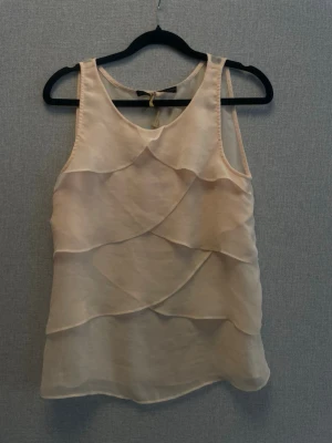 Beige linnetopp med volangdetaljer - Säljer en ljusbeige ärmlös linnetopp med flera lager av volangdetaljer framtill. Toppen har en mjuk, flowy känsla och v-ringad hals. Perfekt för dig som gillar en feminin och luftig stil.