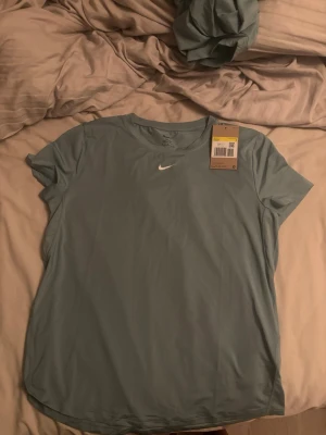 Ljusblå/turkos träningströja från Nike - Helt ny Snygg ljusblå träningströja från Nike med korta ärmar och rund hals. Tillverkad i ett lätt och snabbtorkande material som passar perfekt till gymmet eller löprundan. Diskret Nike-logga på bröstet för en sportig look.