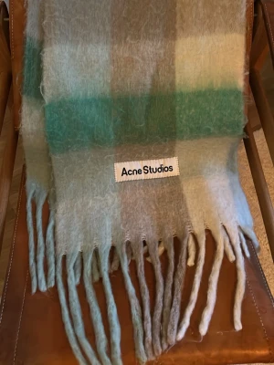 Rutig mohairhalsduk Acne Studios - Säljer en fluffig och mjuk halsduk från Acne Studios i rutigt mönster med nyanser av grönt, grått och vitt. Halsduken har långa fransar i ändarna och Acne Studios-logga framtill. Perfekt för dig som vill ha en trendig accessoar med lyxig känsla.
