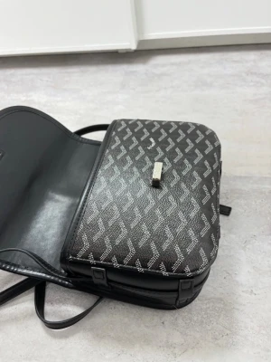 Svart axelväska - (Kan förhandlas vid snabbköpk)Snygg svart axelväska från … med vitmönstrat grafiskt tryck och gul insida. Väskan har justerbar axelrem och stängs med ett metallspänne framtill. Materialet är konstläder och väskan har en klassisk, rektangulär form.