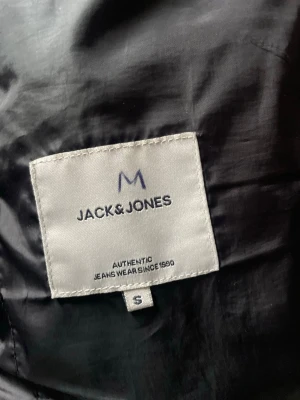 Svart dunväst från Jack & Jones - Snygg svart dunväst från Jack & Jones med quiltad design och diskret logga på bröstet. Västen har en modern look och passar perfekt till lager-på-lager-stil. Tillverkad i syntetmaterial för lätt och bekväm känsla.