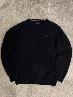 Svart stickad tröja från Gant - Klassisk svart stickad tröja från Gant med rund halsringning och diskret logga broderad på bröstet. Tröjan har långa ärmar och ribbade muddar vid ärmslut och nederkant. Perfekt för lager på lager och enkel att matcha.