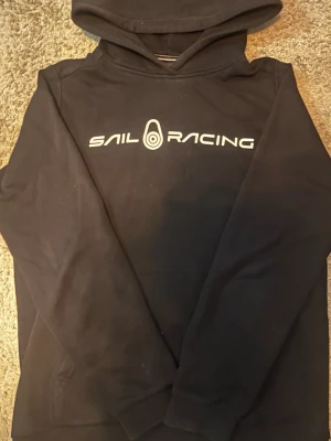 Svart hoodie från Sail Racing - Svart hoodie från Sail Racing med vit logga och text på bröstet. Klassisk modell med huva och ribbade muddar. Tillverkad i mjukt bomullsmaterial som är skönt mot huden. Perfekt för dig som gillar sportig och stilren look.