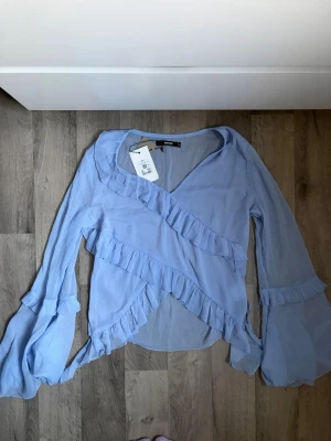 Ljusblå volangblus från Bik Bok - Supersöt ljusblå blus från Bik Bok med omlottmodell och volangdetaljer längs ringningen och framtill. Blusen har långa, luftiga ärmar med volanger vid ärmsluten och är tillverkad i ett tunt, skirt tyg som ger en romantisk vibe. Helt ny, nypris 299kr, se lapp