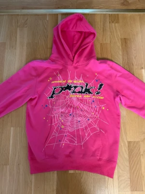 Rosa hoodie med spindelnätsmönster - Cool rosa hoodie med stor huva och tryck framtill. Motivet består av ett vitt spindelnät, färgglada stjärnor och texten 'p*nk!' i svart/vitt med gula detaljer. Tillverkad i mjukt bomullsmaterial, perfekt för en avslappnad streetwear-look.