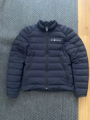 Svart dunjacka från Sail Racing - Snygg svart dunjacka från Sail Racing med diskret logga på bröstet. Jackan har två dragkedjeförsedda fickor fram och quiltad design. Lättviktig och varm med hög krage, perfekt för kalla vinterdagar. Fylld med Allied Feather & Down 800 fill power. FAST PRIS! Allt funkar med den inga fläckar och sådant, dock de ändå är att dragkedjan kan krångla till ibland.