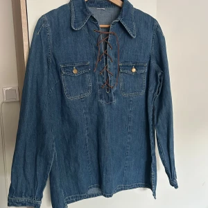 Jeansskjorta med snörning  - Blå jeansskjorta i 100% bomull med cool snörning framtill. Skjortan har två bröstfickor med knappar och klassisk krage. Knappt använd. Osäker på storlek men skulle säga att den passar dem med M. 