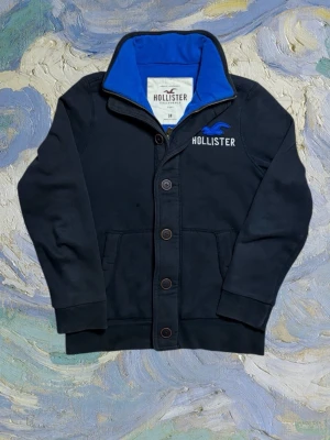 Vintage Hollister cardigan - herr strl M - Säljer en vintage Hollister zip och button up cardigan i svart färg med blå insida i storlek M. Dubbelkrage samt vit “Hollister” brodering på vänster sida av bröstkorgen med blå Hollister bird ovanför. Fickor på båda sidor vid midjan.  👕 Märke: Hollister 📏 Storlek: M 🎨 Färg: Svart med blå insida 🧵 Material: 70% bomull, 30% polyester 📐 Mått: 68 cm lång, 56 cm bred 📌 Skick: Mycket bra skick (se bilder)  💬 Hör av dig vid frågor 💰 Pris kan diskuteras vid snabb affär 📦 Skickas inom 24 timma