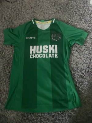 Grön Hammarby matchtröja - Säljer en grön Hammarby fotbollströja från Craft med vita detaljer och trycket 'HUSKI CHOCOLATE' på bröstet. Tröjan har klubbmärke på vänster sida och är tillverkad i ett lätt och ventilerande material, perfekt för träning eller match.