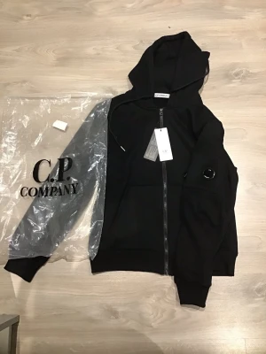 Svart hoodie från C.P. Company - Säljer en svart hoodie från C.P. Company med dragkedja framtill och klassisk linsdetalj på ärmen. Hoodien har fickor på sidorna och justerbar huva. Tillverkad i mjukt material som känns skönt mot huden. Perfekt för dig som gillar streetwear och snygga detaljer.