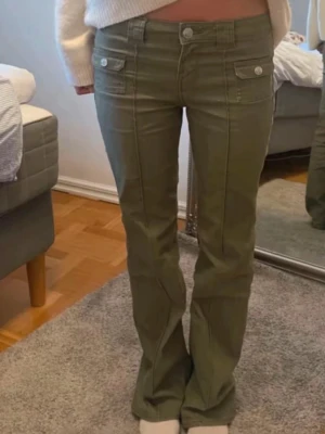 Olivgröna bootcut jeans - Snygga olivgröna jeans med bootcut passform och dubbla fickor framtill med silverfärgade knappar. Jeansen har synliga sömmar och en låg midja som ger en cool retrovibe. Perfekta för dig som vill ha en avslappnad men trendig look. Avklippta längst ner så dom passar någon som är under 165!