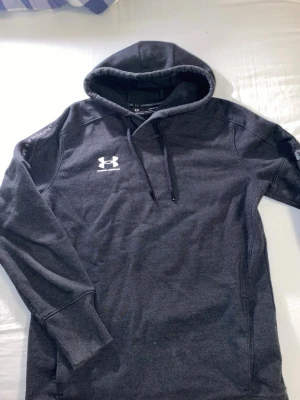 Mörkgrå hoodie från Under Armour - Snygg mörkgrå hoodie från Under Armour med klassisk logga på bröstet. Hoodien har dragsko i huvan och är tillverkad i ett mjukt, fleece-liknande material. Perfekt för chill dagar eller träning. Långa ärmar och ribbade muddar.