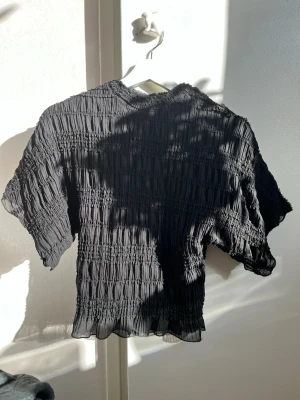 Svart ribbad blus med kort ärm - Trendig svart blus med ribbad struktur och korta ärmar. Toppen har en lätt croppad passform och vågig kant nertill. Perfekt till både jeans och kjol för en cool look.