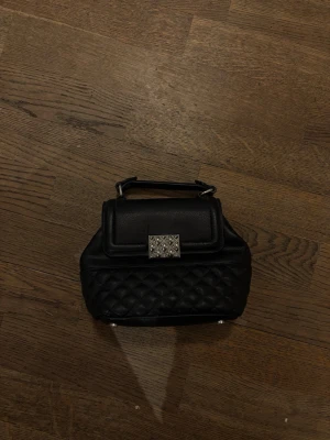 Svart quiltad handväska med metallspänne - Säljer en svart handväska i quiltat konstläder med ett snyggt silverfärgat metallspänne framtill. Väskan har ett kort handtag upptill och en stilren form. 
