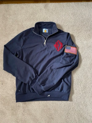 Jeffrey Epstein Halfzip USA Flag 100% Bomull - Marinblå sweatshirt med half zip och hög krage. Broderad röd logga på bröstet och amerikansk flagga på ärmen. Ribbade muddar och nederkant. Perfekt för en sportig och avslappnad stil.