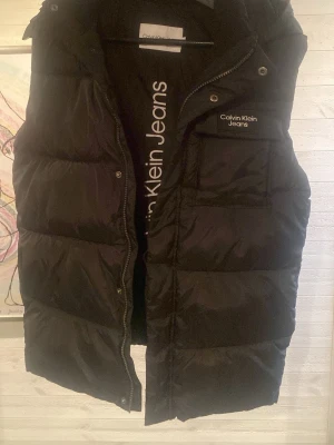 Svart dunväst från Calvin Klein Jeans - Snygg svart dunväst från Calvin Klein Jeans med huva och dragkedja framtill. Västen har broderad logga på bröstet och breda, quiltade paneler för extra värme. Perfekt för lager på lager och streetwear-stil.