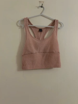 Rosa ribbad linnetopp - Säljer en ljusrosa ribbad linnetopp från H&M. Toppen är croppad och har brottarrygg, vilket ger en sportig vibe. Perfekt att matcha med höga jeans eller shorts. Materialet är mjukt och stretchigt, vilket gör den superbekväm.