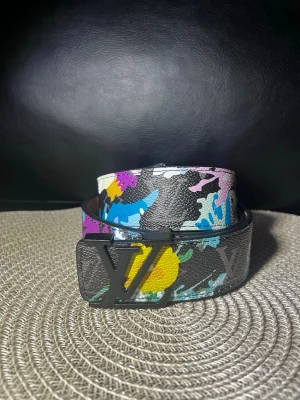 Färgglatt LV-bälte i skinn - Säljer ett Louis Vuitton-bälte i svart skinn med färgglatt kamouflagemönster i blått, gult, rosa och vitt. Bältet har en stor LV-logga i metall som spänne och är tillverkat i Spanien. Perfekt accessoar för att lyfta din outfit.