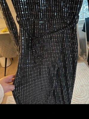 Svart glittrig långklänning - Säljer en svart långklänning med glittriga paljettdetaljer i ränder. Klänningen har en slits på sidan som ger en snygg touch. Perfekt för dig som vill sticka ut med lite extra glitter.