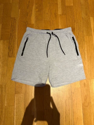 Ljusgrå shorts med snörning & dragkedjor - Säljer ett par ljusgrå shorts från Jack and Jones  med svarta snören i midjan och svarta dragkedjor på fickorna. Mjukt material, perfekt för chill eller träning. Liten logga på vänster ben. 