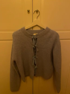 Ljusblå stickad kofta med rosetter - Supersöt ljusblå stickad kofta med långa ärmar och unika rosettdetaljer framtill istället för knappar. Koftan har rund halsringning och är gjord i ett mjukt, ribbstickat material som känns både trendigt och bekvämt.