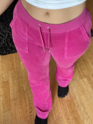 Rosa velour mjukisbyxor med snörning - Säljer ett par snygga rosa juicy mjukisbyxor i mjuk velour med bred resår i midjan och snörning framtill. Byxorna har raka ben och två fickor fram. Perfekta för chill dagar eller när du vill vara extra bekväm.