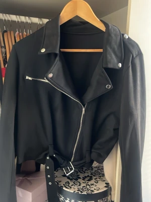 Svart biker jacka i mocca  - Snygg svart mocca jacka  i klassisk bikerstil med silvriga dragkedjor, tryckknappar och axelklaffar. Jackan har ett bälte i midjan och breda slag. Perfekt för en cool och edgy look.