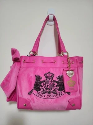 Juicy Couture daydreamer rosa handväska i velour - Superfin rosa handväska från Juicy Couture i mjuk velour med detaljer i konstläder. Väskan har svarta broderier med logga, guldfärgade metalldetaljer och en dekorativ tag med hjärta och krona. Snygg rosett på sidan och praktiska handtag för enkel bärning.