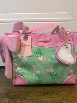 Juicy Couture daydreamer rosa och grön tote - Supergullig handväska från Juicy Couture i rosa och grönt med körsbärsmönster och logga. Väskan har mjukt tyg, rosa detaljer i mockaimitation, rosett på sidan och ett hjärtformat nyckelband. Perfekt för dig som gillar Y2K-stil och färgglada accessoarer.