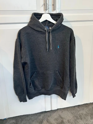 Mörkgrå hoodie från Polo Ralph Lauren - Mörkgrå hoodie från Polo Ralph Lauren med klassisk huva och dragsko. (Gammal modell)Har en känguruficka framtill och den ikoniska blå Polo-loggan broderad på bröstet. Perfekt för en avslappnad och stilren look. Har en defekt vilket inte är svårt o fixa för någon som har möjlighet. Annars bra skick. Kan behöva strykas. Ställ gärna frågor👋😃