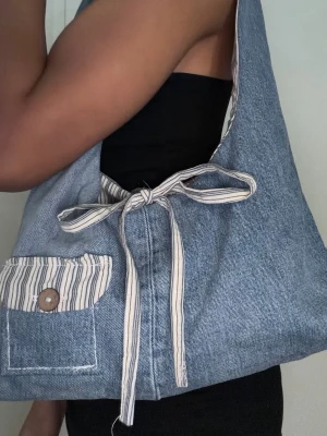 Blå denim handväska - Unik handväska i blå denim. Väskan har en mjuk, avslappnad form och randiga detaljer i vitt och beige. Perfekt för dig som gillar en personlig och trendig stil.