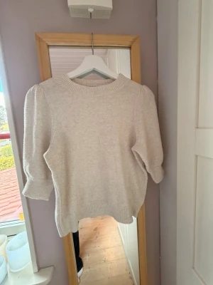Beige stickad tröja med puffärm - Säljer en beige stickad tröja med rund halsringning och trendiga puffärmar. Tröjan har ribbstickade muddar vid ärmslut och nederkant. Perfekt för dig som gillar en stilren och enkel look.