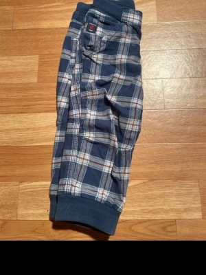 Rutiga blå pyjamasbyxor - Mysiga pyjamasbyxor i blått med rutigt mönster i vitt och rött. Byxorna har muddar vid bensluten, resår i midjan och flera fickor. Perfekta för chillkvällar hemma. Mjukt och bekvämt material.