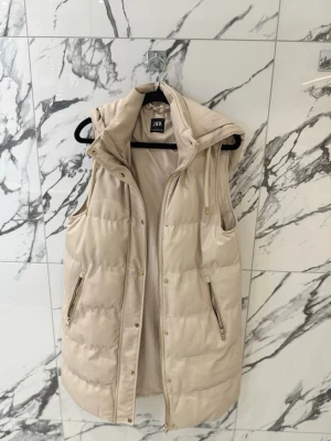 Väst - Zara - Stilren beige dunväst från Zara med huva och dragkedja framtill. Västen är lång, har två fickor med dragkedja och quiltad design. 