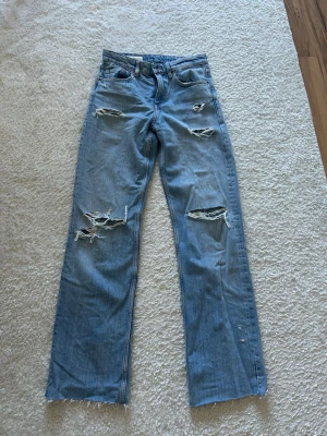 Ljusa slitna jeans från Zara - Säljer ett par ljusblå jeans från Zara med flera slitningar och hål på benen för en trendig, avslappnad look. Modellen är rak med mid waist-låg midja. Aldrig använda då de är för långa för mig