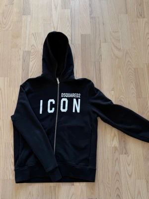 Svart hoodie från Dsquared2 ICON - Svart hoodie med dragkedja från Dsquared2 med stor vit 'ICON'-text och logga på bröstet. Klassisk modell med huva och fickor framtill. Perfekt för en streetwear-look och enkel att matcha med jeans eller joggers. Ny pris 2500-3000 inget kvitto