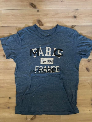 Blå Marc Jacobs t-shirt med tryck - Snygg blå t-shirt från Marc Jacobs med coolt tryck framtill där det står 'Marc Jacobs France Est. 1984'. T-shirten har rund hals och korta ärmar, perfekt för en avslappnad stil. Materialet känns mjukt och skönt mot huden.