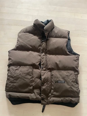 Canada Goose väst - Snygg brun dunväst från Canada Goose med hög krage och stora framfickor. Västen har en klassisk puffer-design och stängs med både dragkedja och tryckknappar. Perfekt för lager-på-lager och håller dig varm under kyliga dagar.