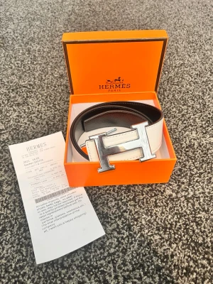  H-bälte i svart och vitt läder - Snyggt H-bälte i svart och vitt läder med den ikoniska H-spännet i silver. Kommer i original orange H-box och med kvitto. Perfekt accessoar för att lyfta vilken outfit som helst med exklusiv känsla.