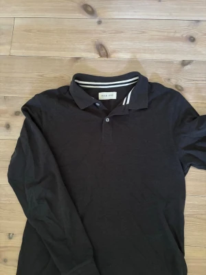 Grå långärmad polotopp  - Svart långärmad polotopp från Pier One med klassisk krage och knappslå. Kragen har en vit randig detalj på insidan som ger en sportig touch. Perfekt för en stilren och avslappnad look.