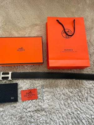 Svart skärp med H-spänne - Säljer ett svart skärp i skinn med ett H-spänne i silver. Bältet har vit kontrastsöm och kommer med originalförpackning, påse och certifikat. Perfekt accessoar för att lyfta vilken outfit som helst. Bältet är 110 cm och priset kan diskuteras