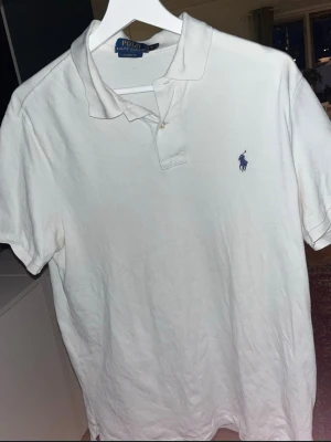 Vit pikétröja från Ralph Lauren - Klassisk vit pikétröja från Ralph Lauren med blå broderad logga på bröstet. Tröjan har krage, knappar vid halsen och korta ärmar. Perfekt till jeans eller shorts för en clean och stilren look.