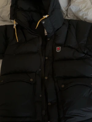 Svart dunjacka från Fjällräven - Säljer en svart dunjacka från Fjällräven med stor huva och gula dragskor. Jackan har två stora framfickor med knappar och klassisk Fjällräven-logga på bröstet. Perfekt för kalla vinterdagar och riktigt varm tack vare dunfyllningen.