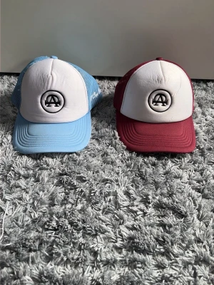 All cap one kepsar - Säljer två snygga kepsar med vit frontpanel och broderad logga. Ena kepsen har ljusblå skärm och mesh, den andra har vinröd skärm och mesh. Klassisk trucker-modell med justerbar snapback och böjd skärm. 200 för båda