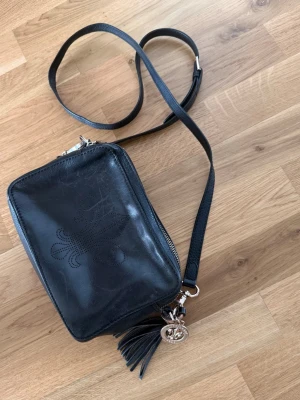 Svart axelväska från Morris Lady i äkta läder - Snygg svart axelväska i läder från Morris Lady i äkta läder med perforerat logomönster på framsidan. Väskan har justerbar axelrem, dragkedja upptill och dekorativ tofs med guldfärgad logodetalj. Perfekt för att bära det viktigaste med stil.