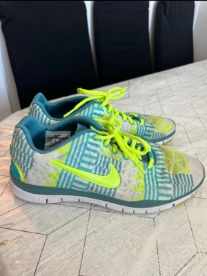 Nike sneakers med neonmönster - Nike sneakers med vit bas, turkosa och neongula grafiska mönster samt neongula skosnören och swoosh. Skorna har lågt skaft och vit sula med turkosa och gula detaljer undertill. Perfekta för dig som gillar färg och sportig stil.