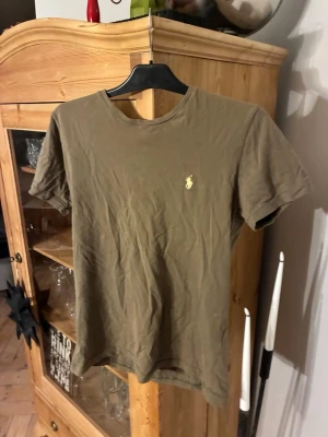 Olivgrön t-shirt från Polo Ralph Lauren - Olivgrön t-shirt från Polo Ralph Lauren med klassisk rund hals och korta ärmar. T-shirten har en broderad gul logga på bröstet och är tillverkad i mjuk bomull. Perfekt för en avslappnad och stilren look. Använd ett fåtal gånger och har storleken M